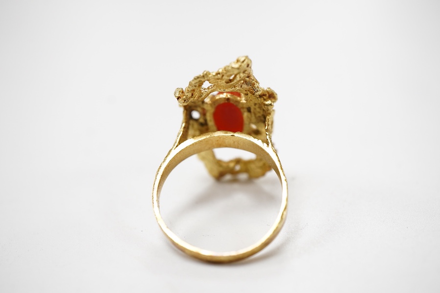 A gold and coral ring, 1970s, of textured design, set with a coral cabochon, mounted in gold, size N1/2, stamped 18K, gross weight approximately 7 grams